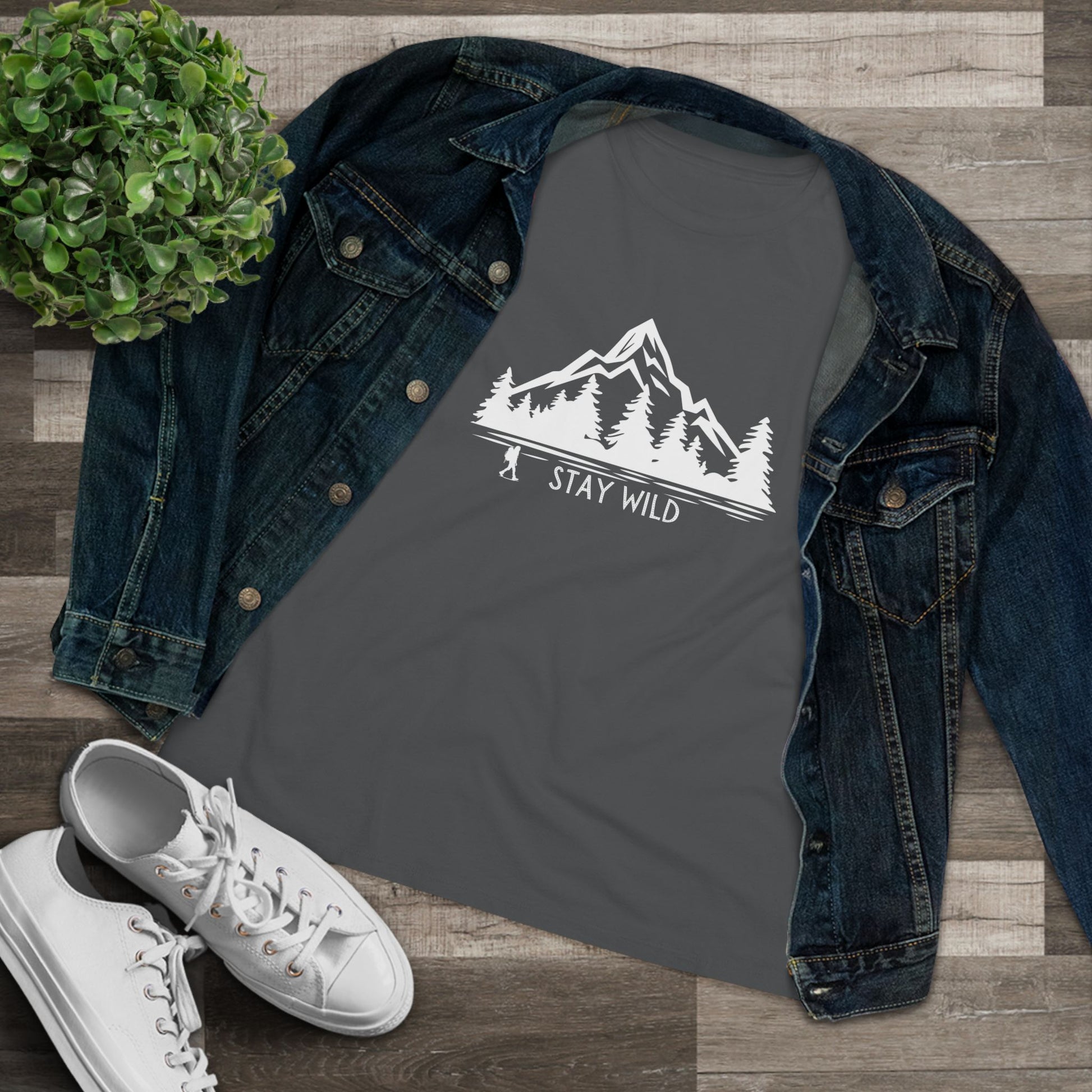 Dark gray t-shirt with 'Stay Wild' graphic on a denim jacket and white sneakers, on a wooden surface.